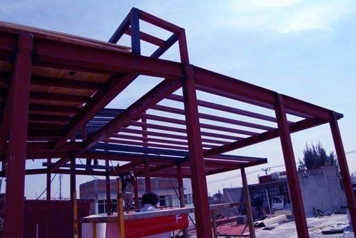 Estructura de acero roja de un edificio en construcción, con vigas horizontales que forman un techo y un cielo parcialmente nublado en lo alto.