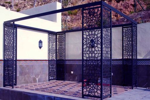 Un cenador de metal oscuro con intrincados paneles cortados a láser. Se alza sobre una plataforma de azulejos, probablemente un espacio de oración al aire libre, con paredes blancas al fondo.