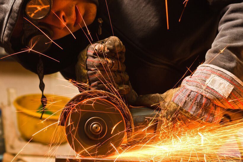Persona que usa gafas de seguridad y guantes moliendo metal, generando chispas en un taller.