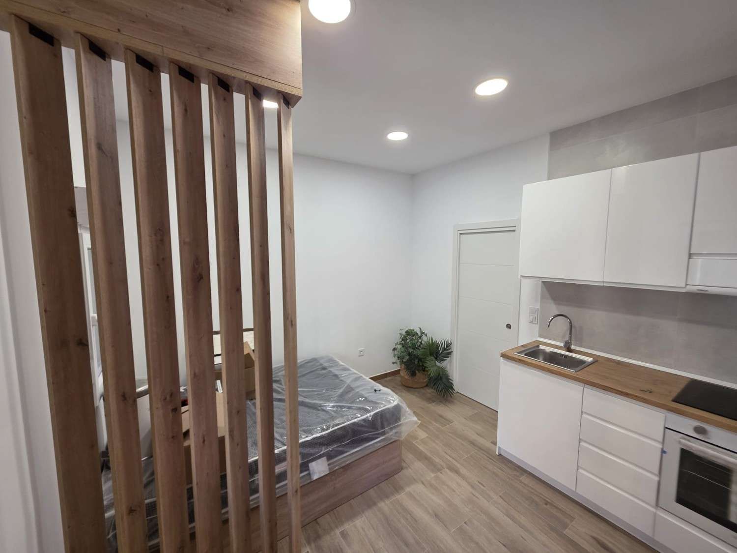 Pequeña zona de cocina con armarios blancos y un separador de ambientes de madera, junto a una cama con colchón gris.