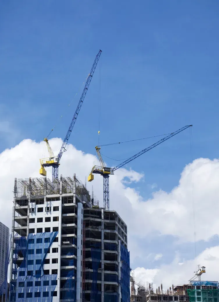 Dos grúas torre se alzan sobre un rascacielos en construcción, con un cielo azul nublado de fondo.