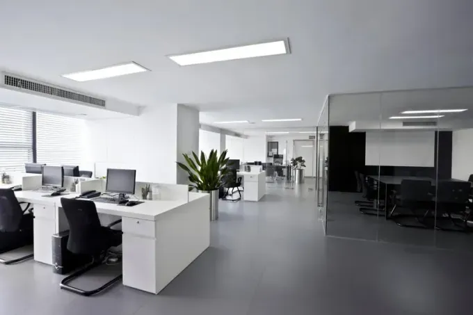Un espacio de oficina luminoso y minimalista con escritorios blancos, sillas negras y una sala de conferencias acristalada.