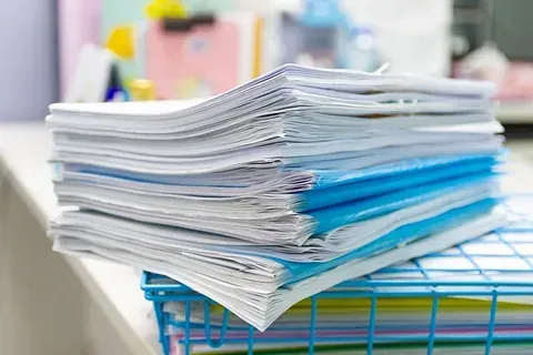 Una pila de papeles reposa sobre una cesta de alambre en un escritorio.