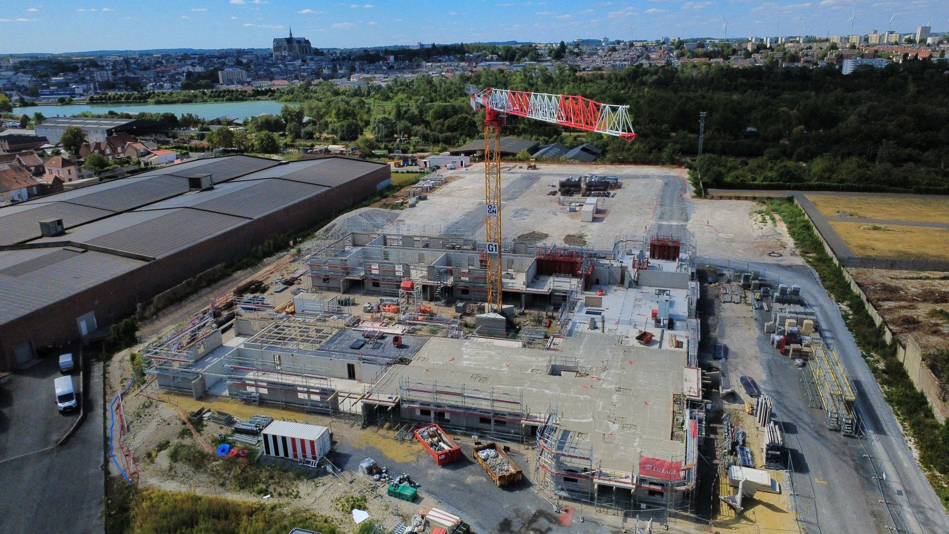 Vue aérienne d'un grand chantier de construction avec des grues, des structures en béton, et des routes et des cours d'eau à proximité.