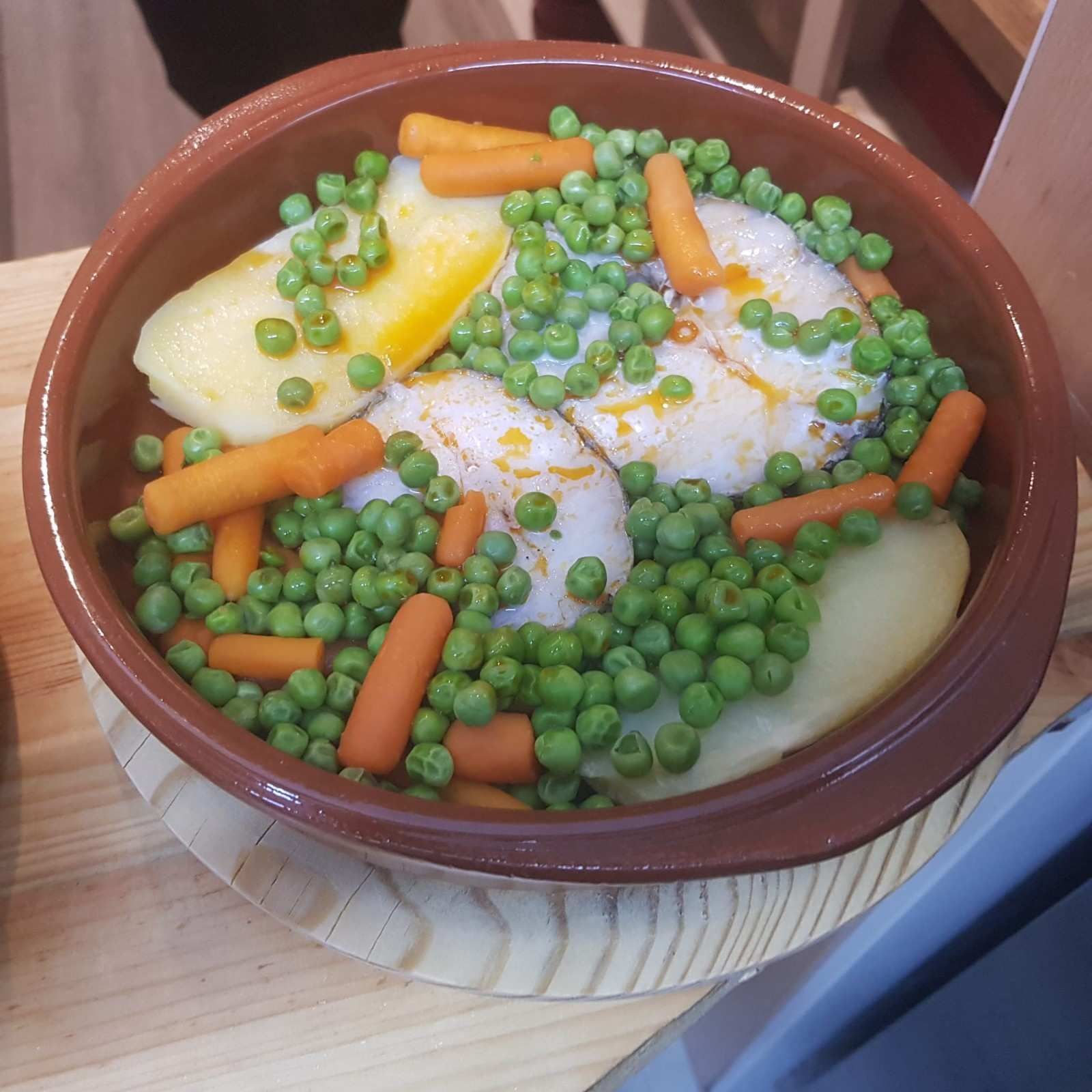 Un plato de barro marrón contiene filetes de pescado blanco, patatas cocidas, zanahorias baby y guisantes verdes en una salsa sabrosa.