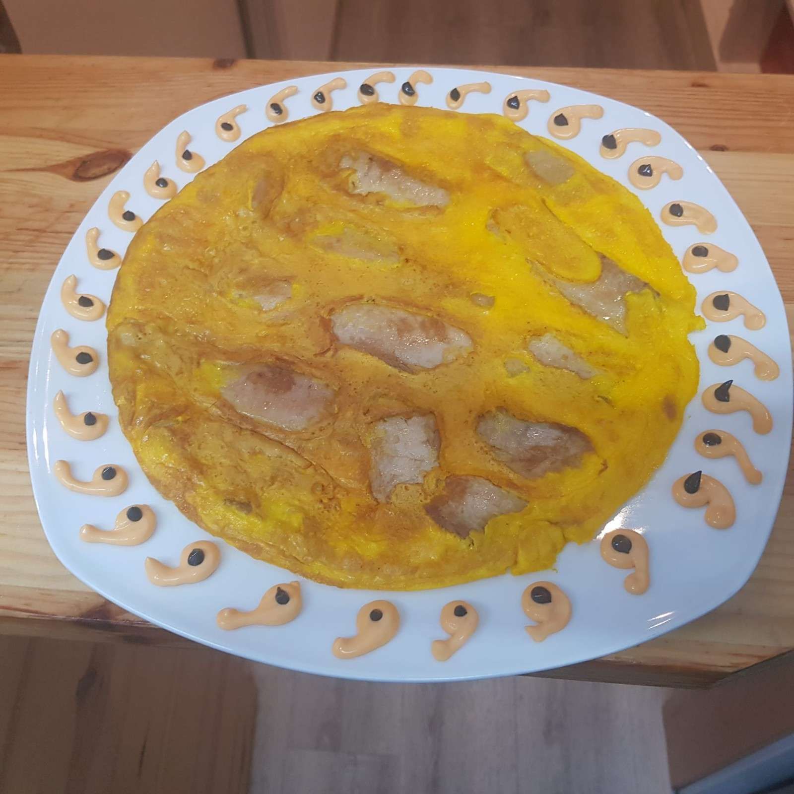 Una tortilla de patatas circular, centrada en un plato blanco y bordeada por un dibujo decorativo de pequeños puntos de salsa y hierbas.