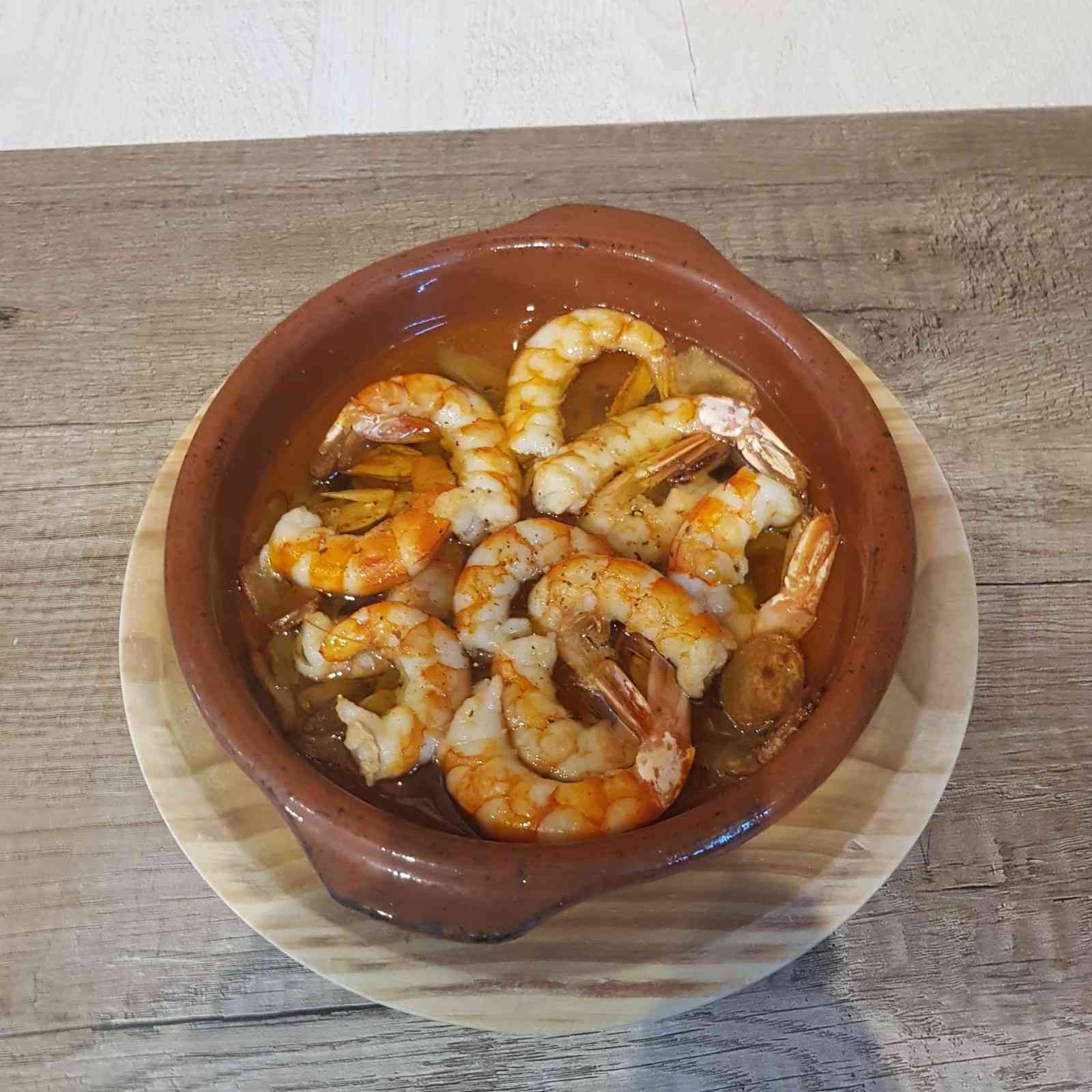 Un pequeño cuenco de cerámica marrón que contiene gambas en una salsa de aceite sazonado, servido sobre una tabla de madera.