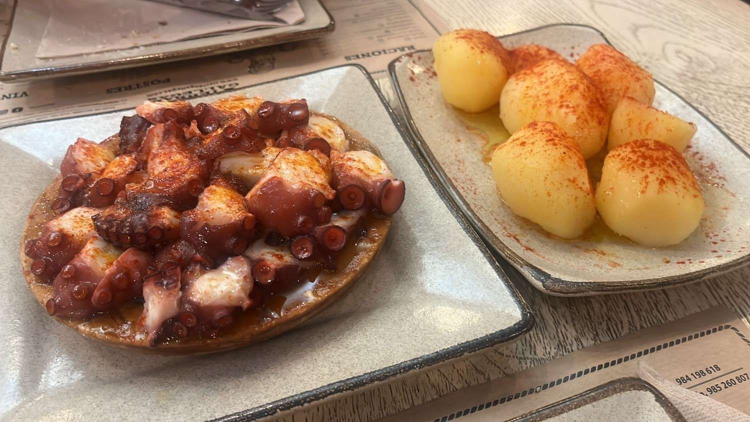 Dos platos blancos sobre una mesa, uno con pulpo y el otro con patatas cocidas sazonadas con pimentón.