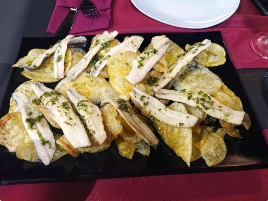 Un plato rectangular negro cubierto con patatas fritas y filetes de anchoa blanca en escabeche, adornado con hierbas aromáticas.