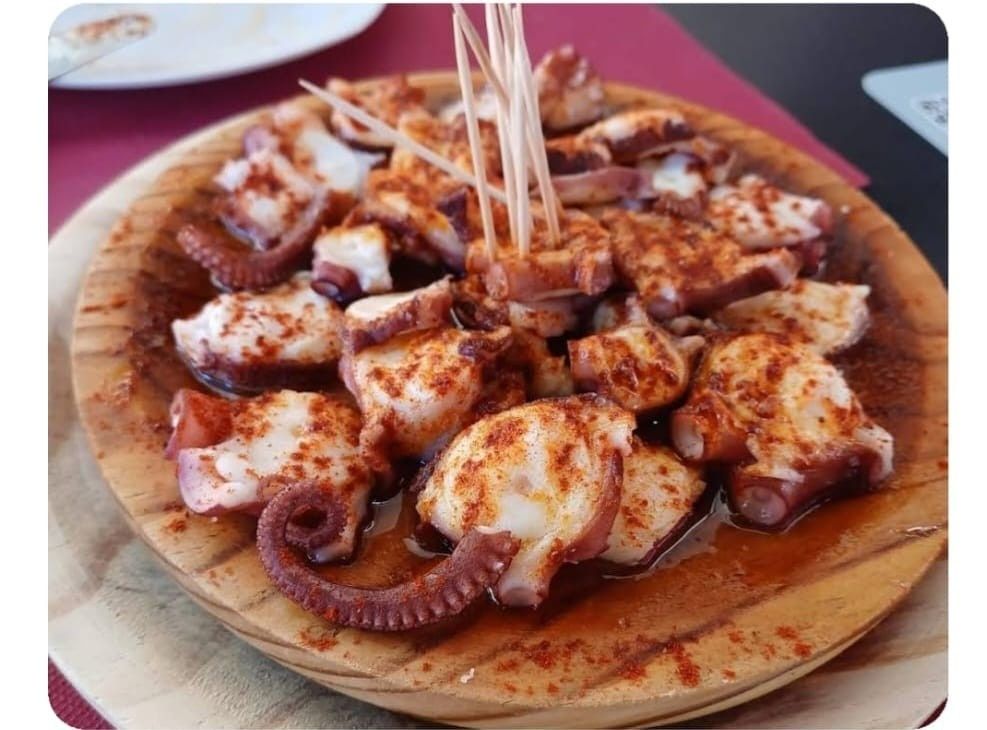 Un plato de madera lleno de pulpo en rodajas, aderezado con pimentón y aceite de oliva, con varios palillos para servir.