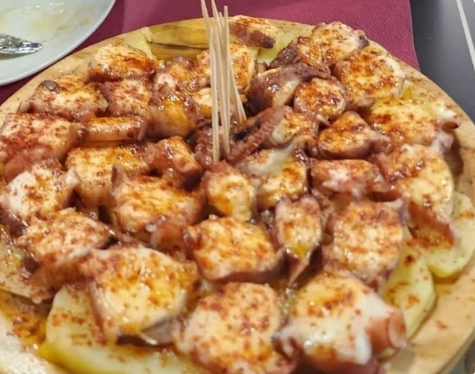 Un plato de madera lleno de pulpo en rodajas, aderezado con pimentón y aceite de oliva, servido sobre una cama de rodajas de patata.