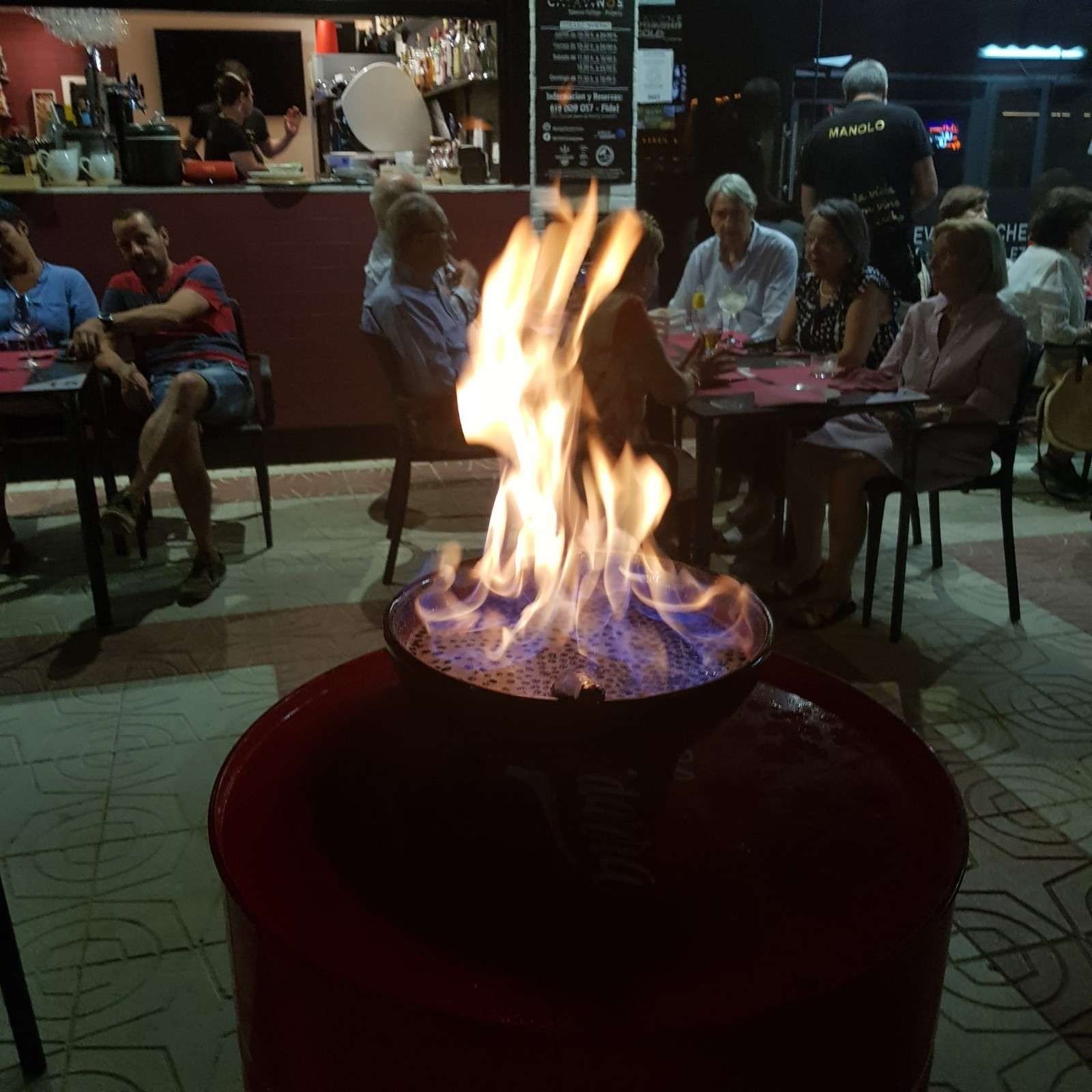 Un fuego brillante arde en un cuenco de metal en primer plano, en la terraza tenuemente iluminada de un restaurante al aire libre con clientes sentados.
