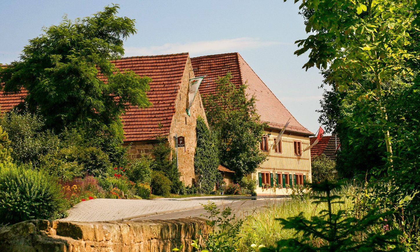 Stein- und gelbes Haus mit rotem Ziegeldach, umgeben von grünen Bäumen und Pflanzen; sonniger Tag.