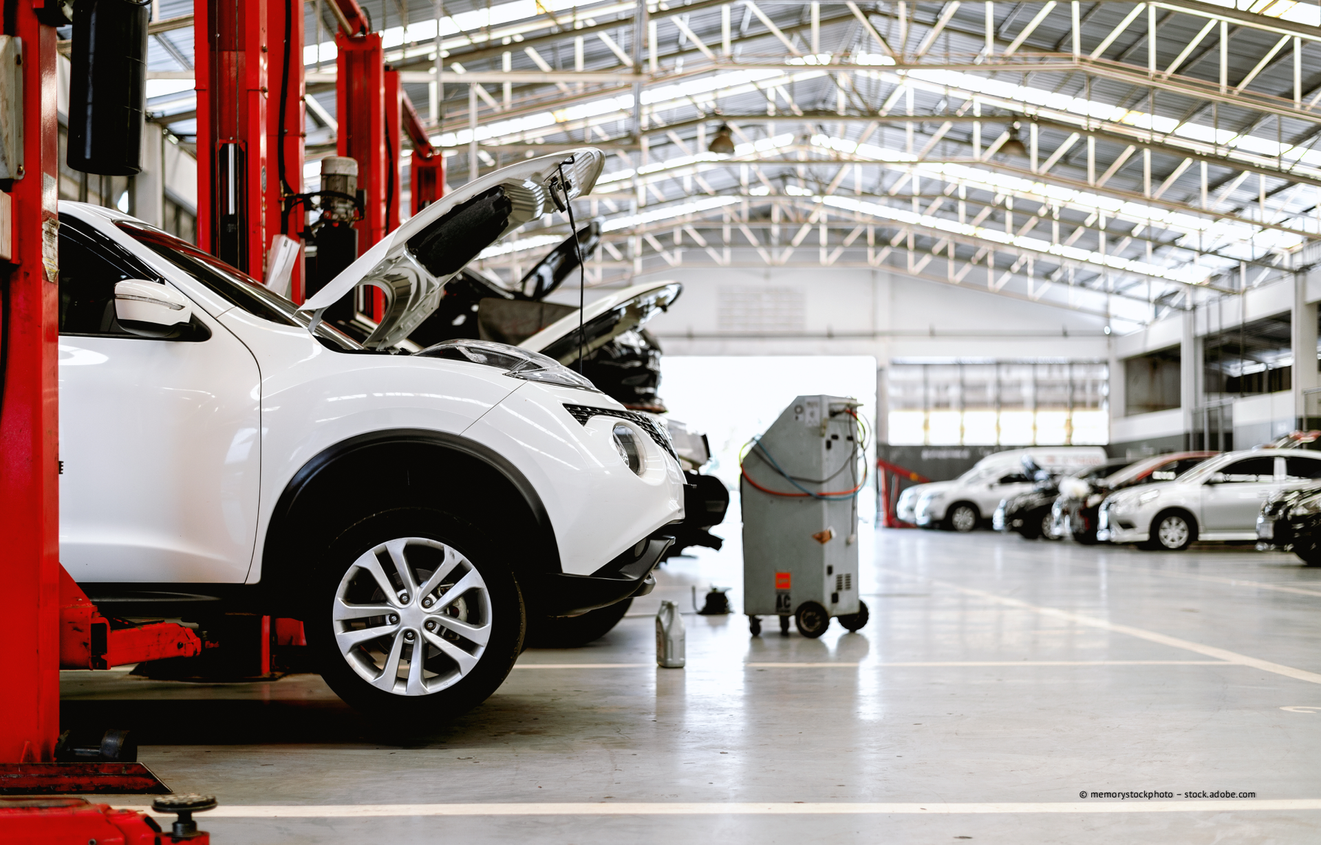 Ein weißes Auto mit geöffneter Motorhaube steht auf einer Hebebühne in einer Autowerkstatt neben einem anderen Fahrzeug und einem Werkstattwagen.