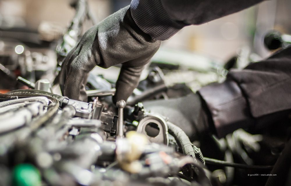 Ein Mechaniker rollt in einer Autowerkstatt einen Autoreifen; im Hintergrund steht ein Fahrzeug auf einer Hebebühne.