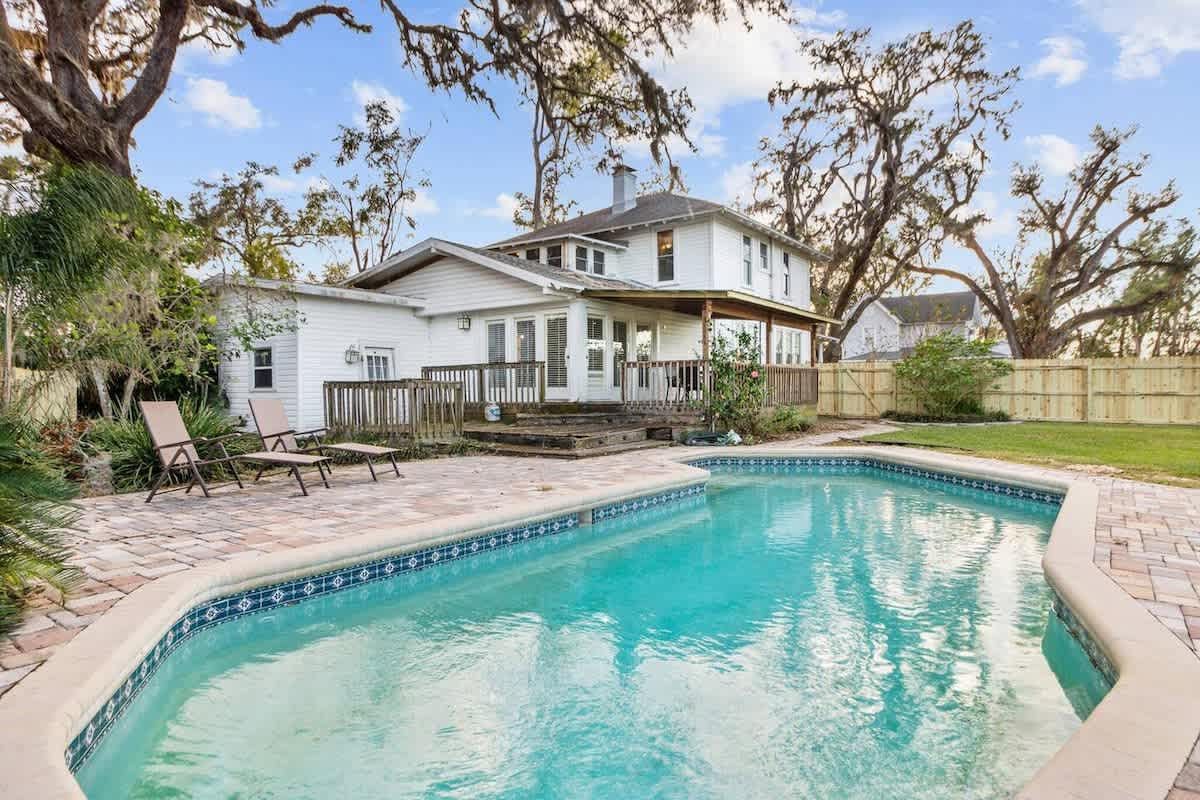 Backyard with pool, white house, wooden deck, green lawn, trees, and blue sky.