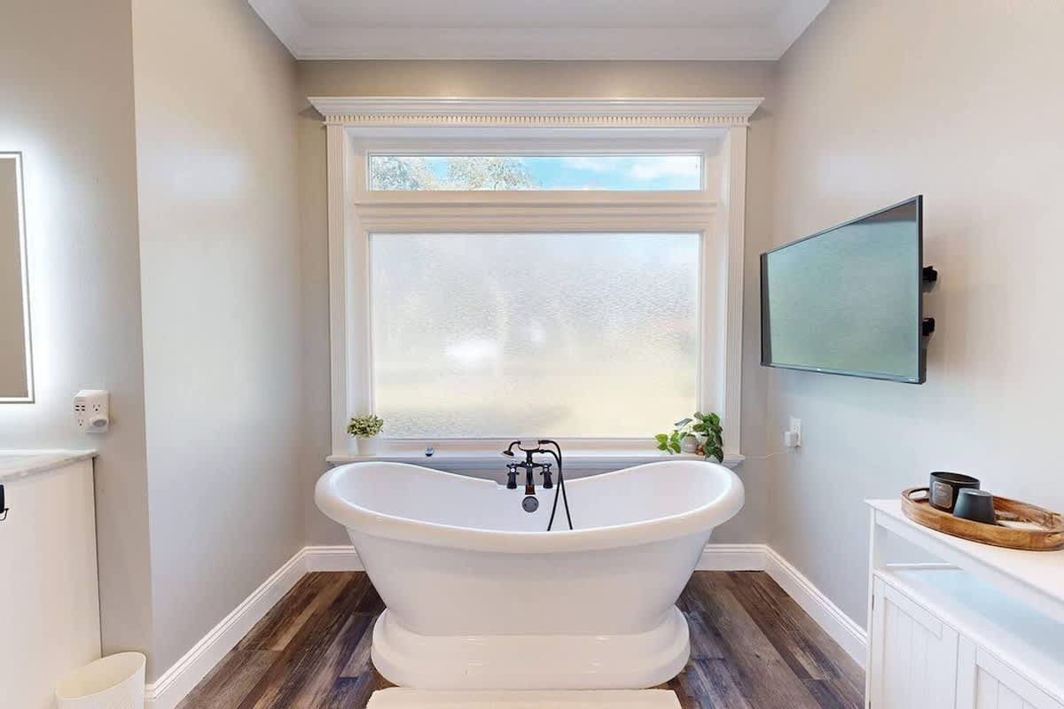 Bathroom with freestanding tub under window, TV on wall, white vanity.