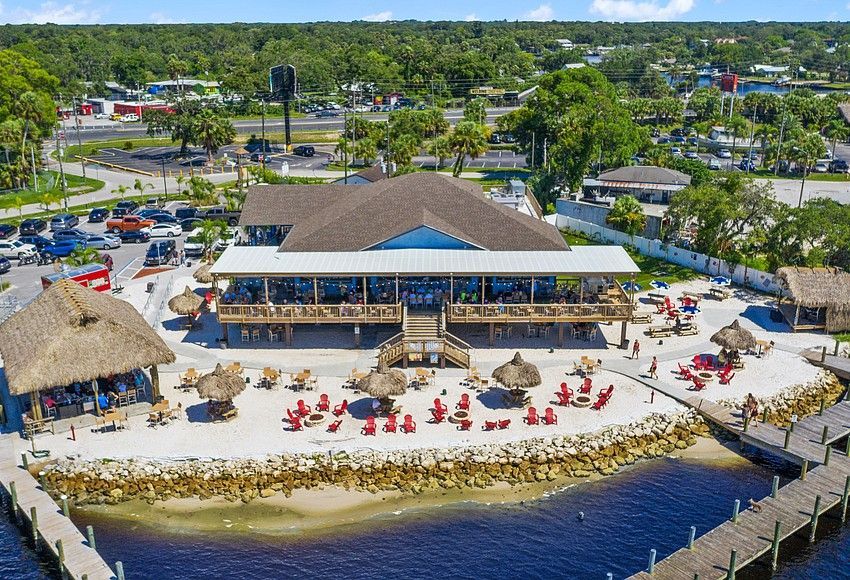 Restaurant with a thatched-roof tiki hut, deck seating, and red chairs on a sand area next to a body of water.