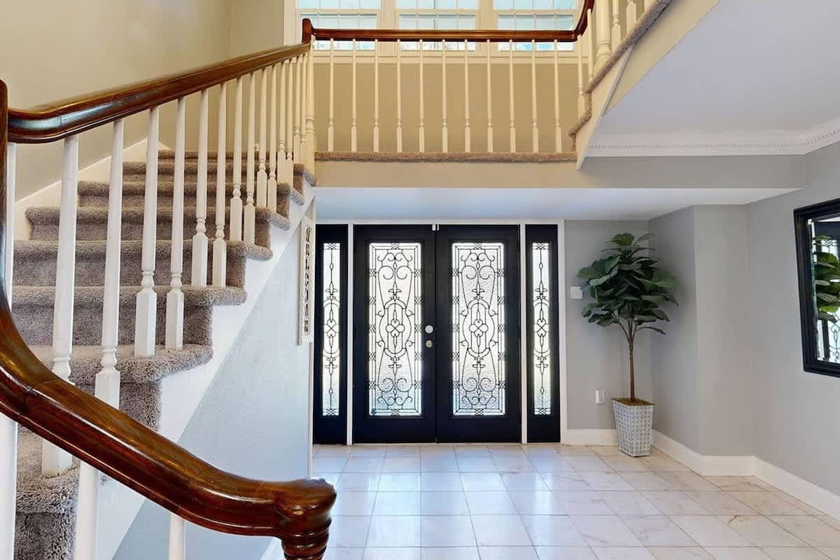 Grand foyer with double glass doors, a staircase with a wooden banister, and a potted tree.
