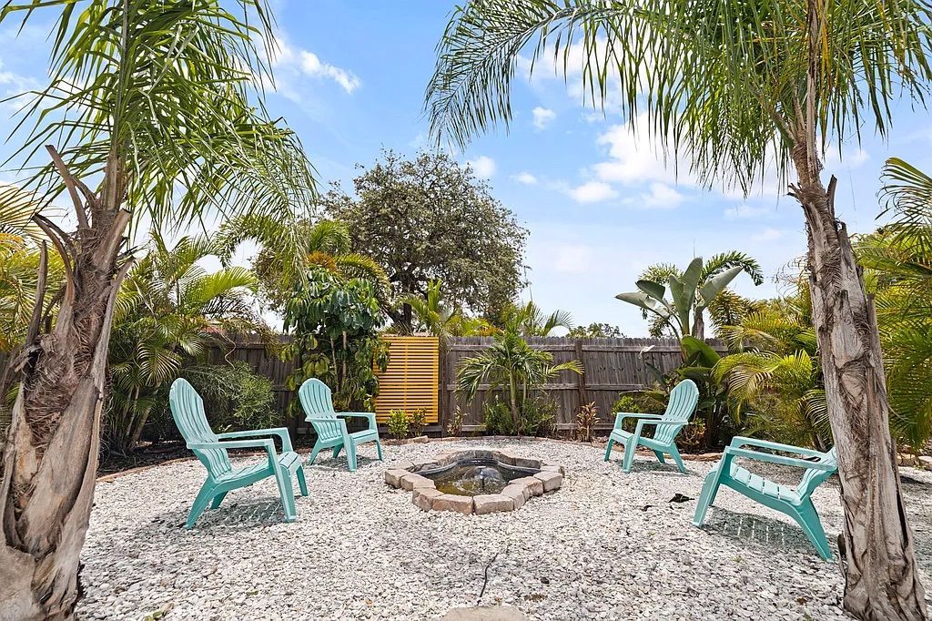 Backyard with white gravel, turquoise chairs around a fire pit, palm trees and greenery.
