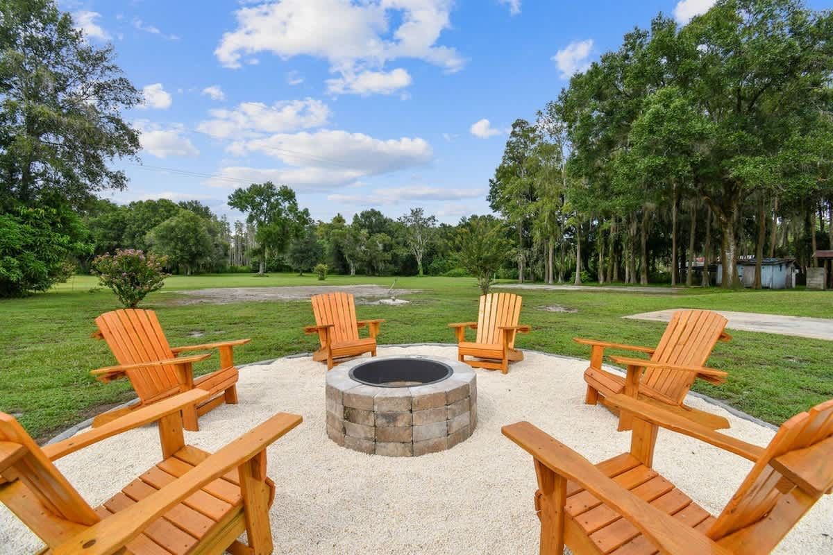 Six wooden chairs around a stone fire pit on a gravel patio, green lawn, trees, blue sky.