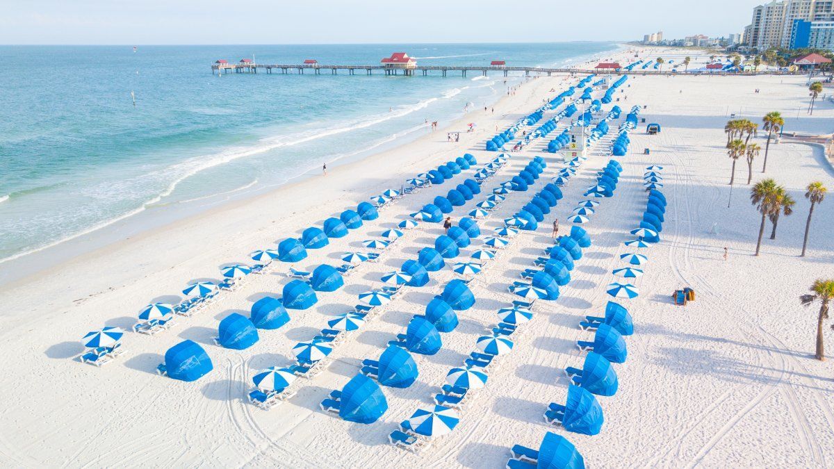 Aerial view of a sunny beach with rows of blue umbrellas and chairs, pier in the distance, and blue ocean.