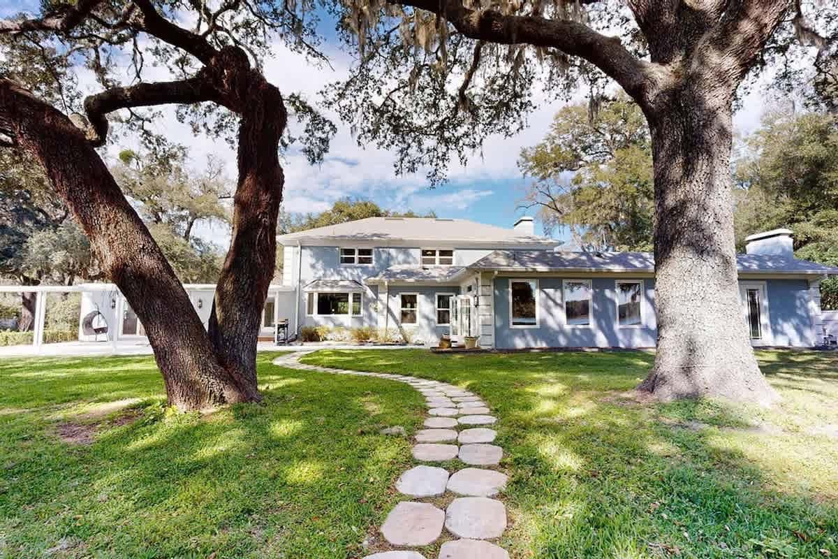 Stone path leading to a two-story blue house, framed by large trees on a green lawn.
