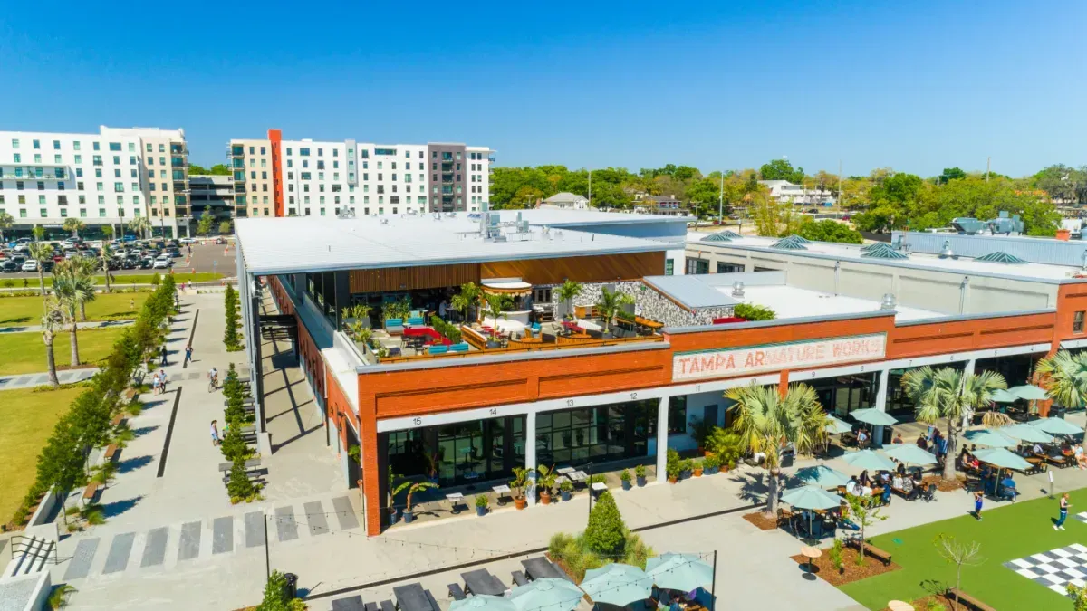 Aerial view of a shopping center with outdoor seating, green space, and apartment buildings in the background under a blue sky.