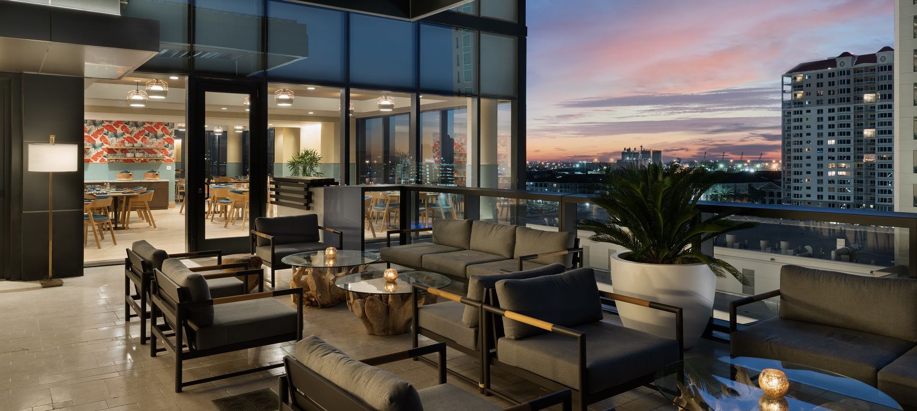 Rooftop lounge at sunset with couches, chairs, small tables, and a city skyline view.