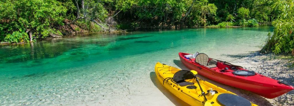 Two kayaks, one yellow and one red, rest on a sandy shore of a clear turquoise river lined with lush green trees.