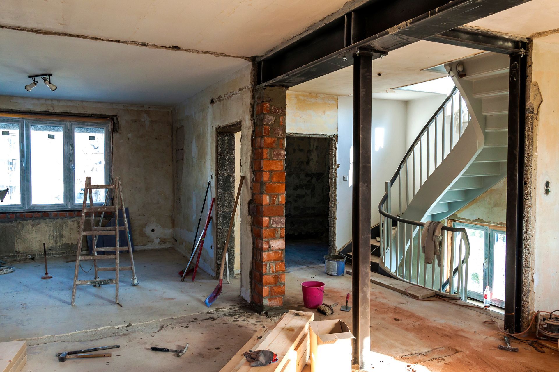 Pièce partiellement rénovée, avec des briques apparentes, une échelle, des outils et un escalier visibles à travers une porte.
