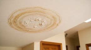 Plafond endommagé par l’eau, avec une grande tache brune et de la peinture qui s’écaille, près d’un couloir et d’une porte en bois.