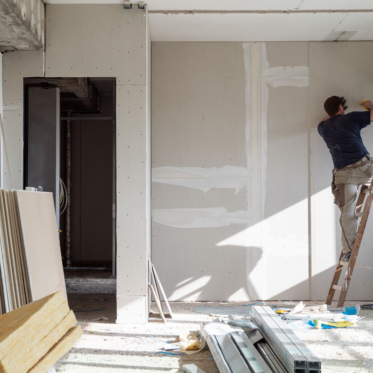 Un ouvrier sur une échelle peint un mur intérieur blanc dans une pièce partiellement rénovée.