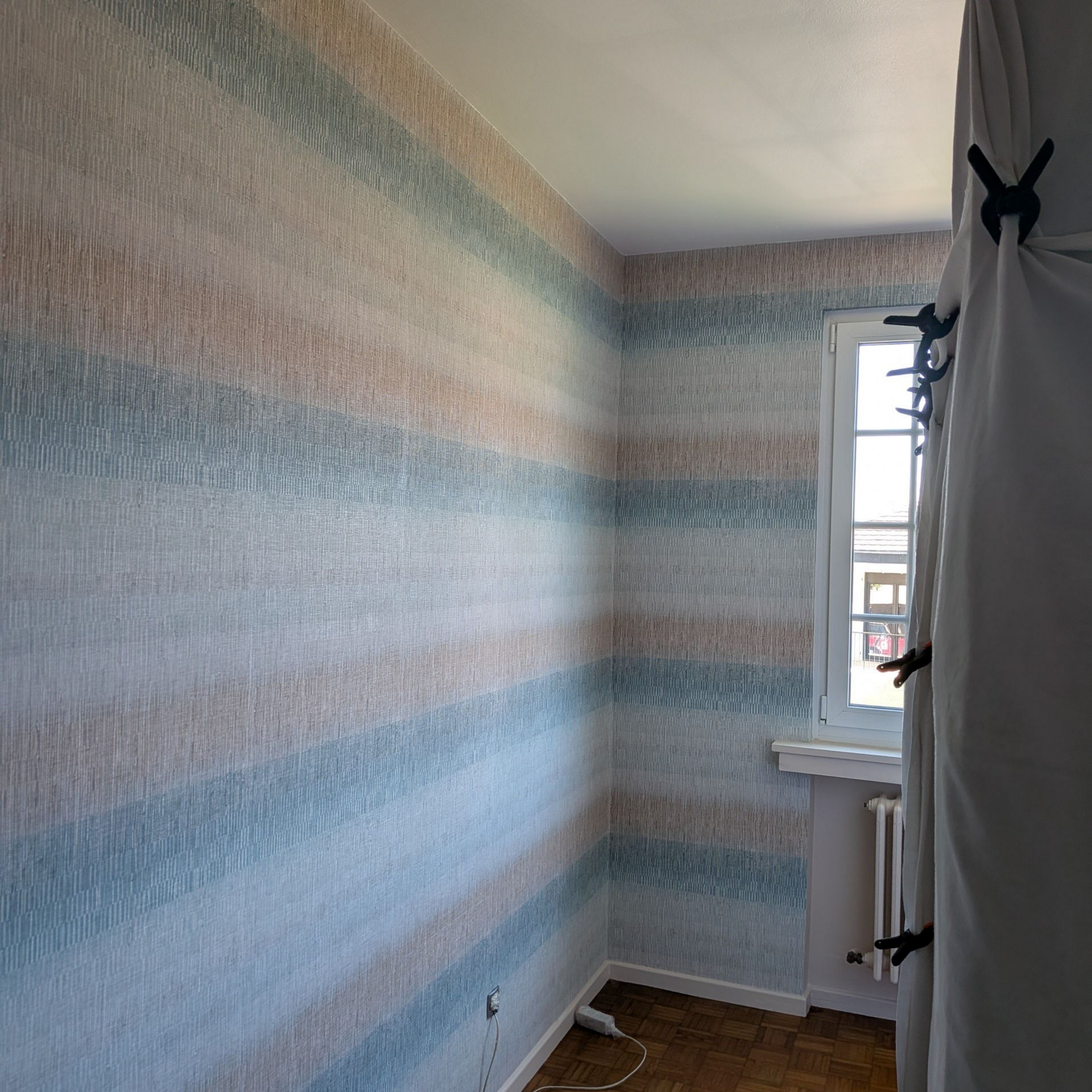 Pièce étroite aux murs rayés bleu-gris, avec une petite fenêtre et un radiateur sous la fenêtre.