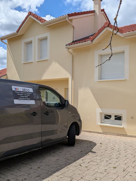 Fourgon sombre garé devant une maison beige au toit de tuiles rouges et aux volets blancs fermés.