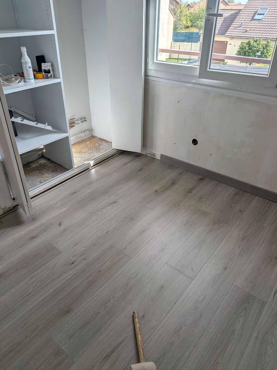 Pièce vide avec un parquet clair, des murs blancs, une fenêtre et une étagère blanche à gauche.