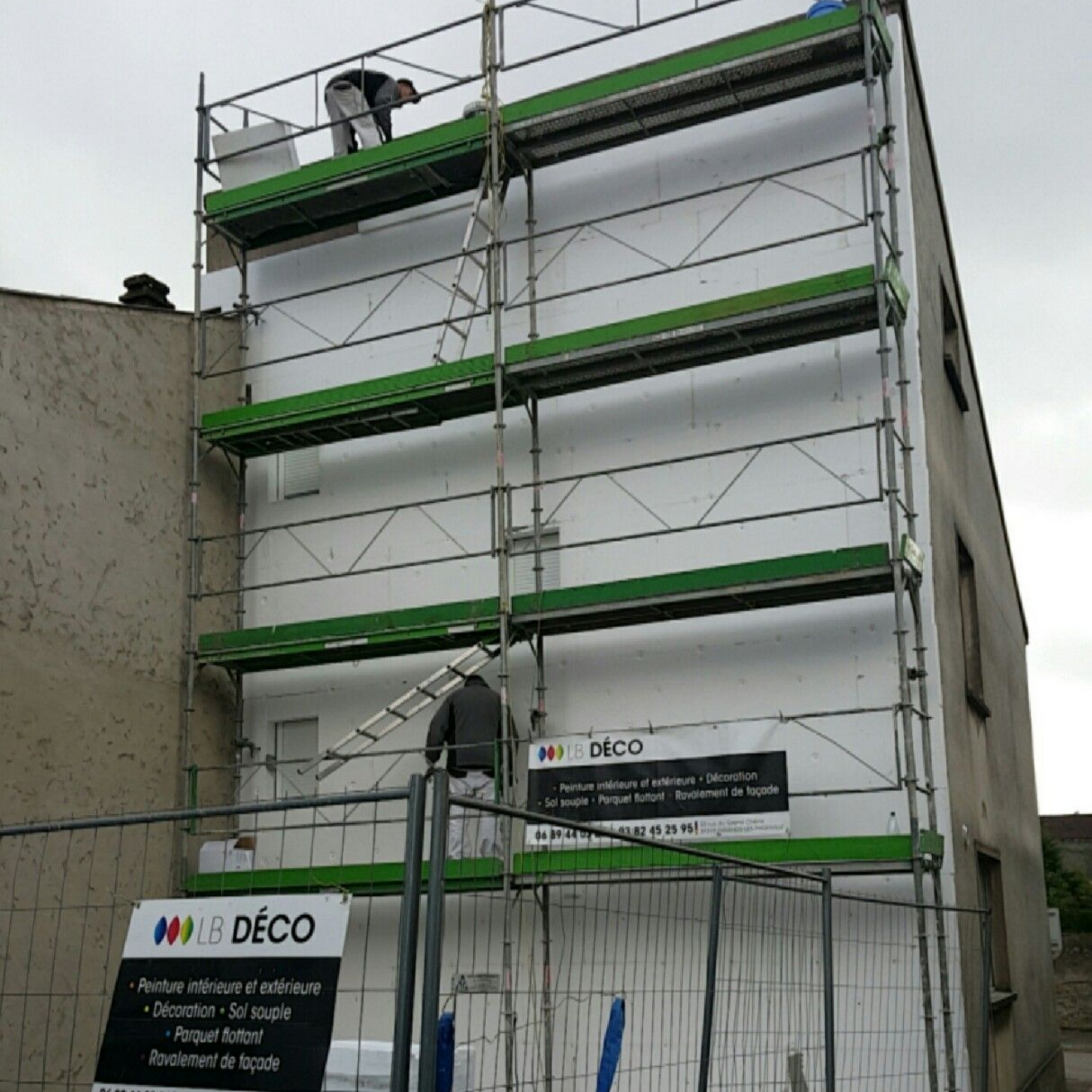 Bâtiment blanc en cours de construction, entouré d’un filet vert, avec une benne bleue et des panneaux de chantier devant.