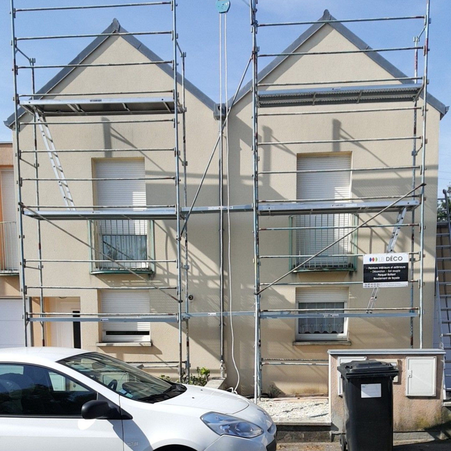 Façade d’une maison mitoyenne entourée d’échafaudages, avec deux voitures garées devant, dont une camionnette blanche, et une poubelle.