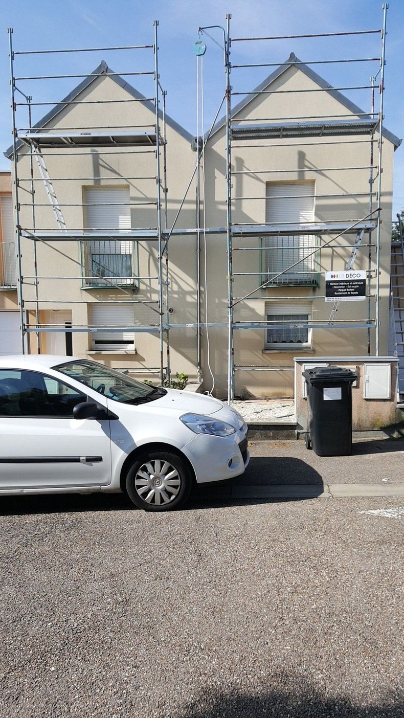 Façade d’une maison de ville en rénovation, entourée d’échafaudages, avec une voiture blanche garée devant.