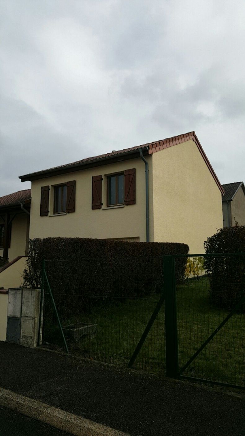 Maison beige à deux étages avec des volets marron, derrière une haie sombre, par temps nuageux.