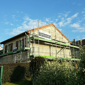 Maison en construction avec des échafaudages et un filet de protection vert sur fond de ciel bleu.