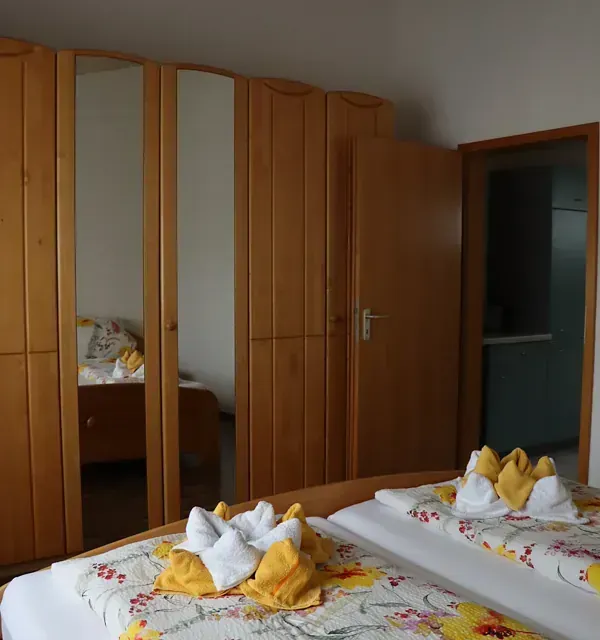 Schlafzimmer mit zwei Betten, geblümter Bettwäsche, Holzschrank, offener Tür zum Badezimmer.
