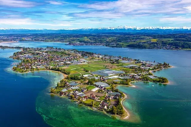 Luftaufnahme einer von blauem Wasser umgebenen Insel mit grüner Vegetation und Gebäuden sowie Bergen in der Ferne.