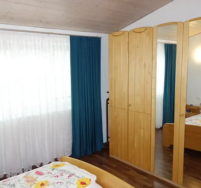 Schlafzimmer mit Holzschrank, Bett und weiß-blauen Vorhängen.