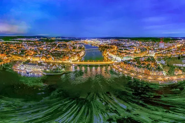 Nächtliche Luftaufnahme einer Stadt mit beleuchteten Gebäuden entlang eines Flusses, mit Brücken und Spiegelungen im Wasser.