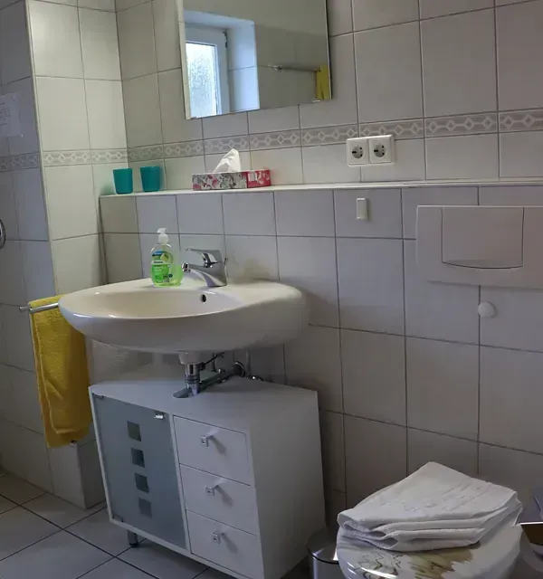 Badezimmer mit weißen Fliesen, Waschbecken und Schrank. Ein Handtuch hängt, die Toilette ist sichtbar.