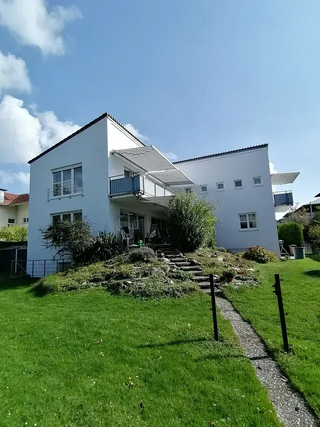 Weißes, modernes Haus mit mehreren Balkonen auf einem grasbewachsenen Hügel unter klarem, blauem Himmel.