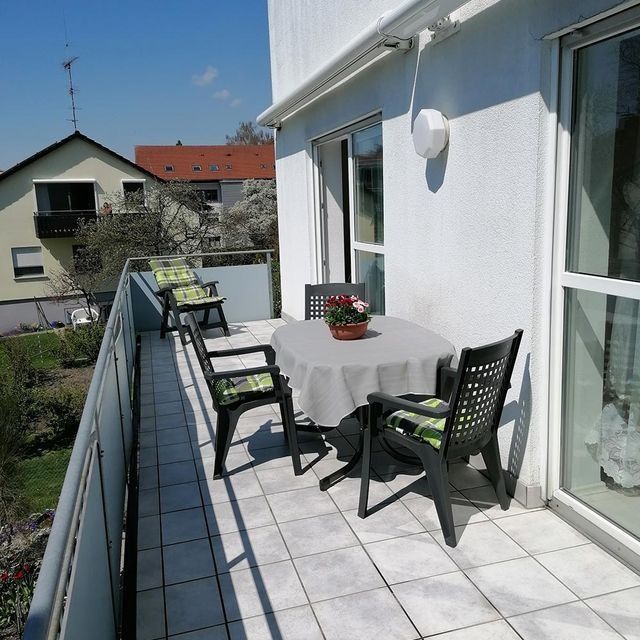 Balkon mit Tisch, Stühlen und Topfblumen. Weiße Wände, Fliesenboden und Blick auf die Häuser.