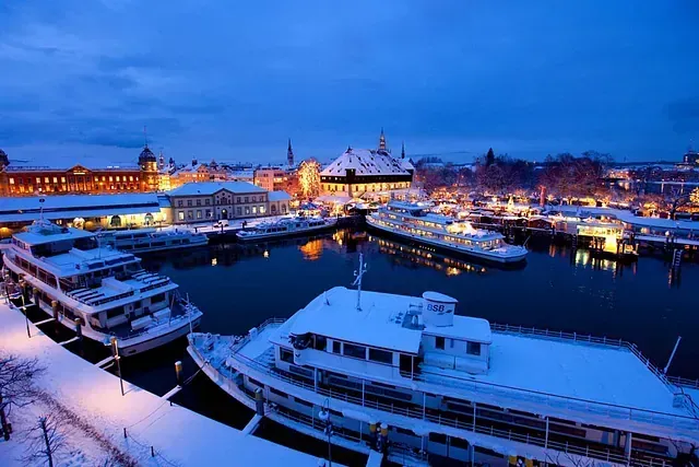 Verschneiter Hafen in der Abenddämmerung, Boote, Gebäude und Stadtlichter spiegeln sich im Wasser.