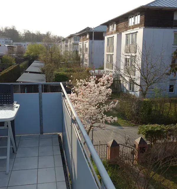 Balkonblick: Wohnhäuser, blühender Baum, grüne Hecken, sonniger Tag.